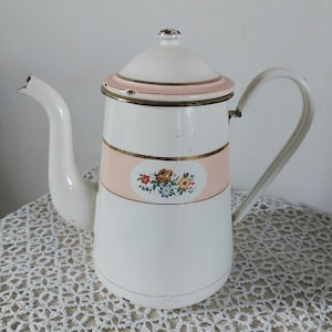 Peut inclure: Cafetière ancienne en émail blanc avec un bec verseur et une poignée incurvés. La cafetière présente une bande rose avec un motif floral et une bordure dorée. Le couvercle et la poignée sont blancs.