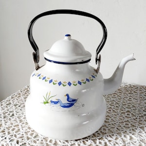 Peut inclure: Théière en émail blanc avec une anse et un bec noirs. La théière présente un motif floral bleu et jaune et une illustration de canard. Le couvercle est blanc avec une bordure bleue. Un article de cuisine vintage.