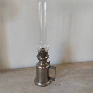 Op de afbeelding: Een zilveren olielamp met een glazen schoorsteen en een handvat. De lamp heeft een decoratief ontwerp rond de basis.