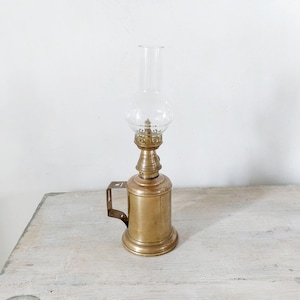 Könnte beinhalten: Eine antike Öllampe aus Messing mit einer Glaskugel und einem Schornstein. Die Lampe hat einen Griff an der Seite und steht auf einer Holzoberfläche. Die Lampe hat eine warme Messingfarbe.