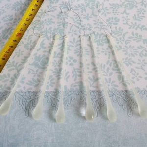 May include: Six clear, teardrop-shaped glass ornaments, each approximately 5 inches long, hang from thin wires. A yellow measuring tape is visible in the upper left corner. The ornaments are arranged on a light blue floral patterned fabric.