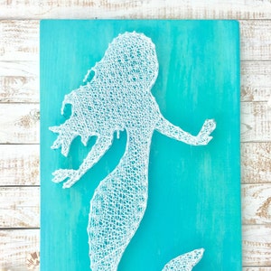 May include: A white string art mermaid silhouette on a turquoise blue wooden background.