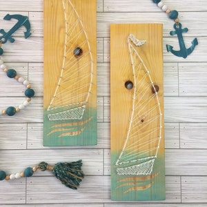 May include: Two wooden sailboat string art pieces. The sailboats are made with white string on a yellow and teal painted wood background. Decorative nautical elements, including an anchor and beads, are also present.