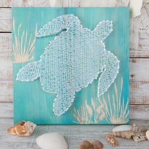 May include: A white string art sea turtle on a turquoise blue wooden background. The turtle is surrounded by white string art seagrass.