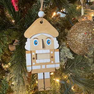 May include: A wooden nutcracker ornament with a white beard, blue eyes, and a brown hat. The ornament is hanging on a Christmas tree.