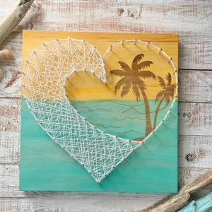 May include: A wooden heart-shaped board with a white string art design of a wave and palm trees. The background is painted in shades of blue and yellow.