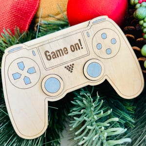 May include: A wooden Christmas ornament shaped like a video game controller. The ornament is light brown and has a blue button design. The text "Game on!" is engraved on the ornament.