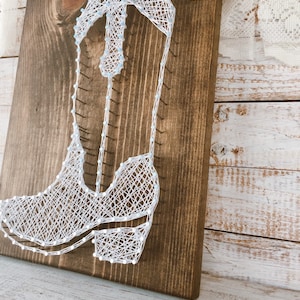 Puede incluir: Arte de hilo blanco de una bota vaquera sobre un fondo de madera marr&oacute;n oscuro. La bota est&aacute; delineada con hilo blanco y los detalles est&aacute;n rellenos con hilo blanco.