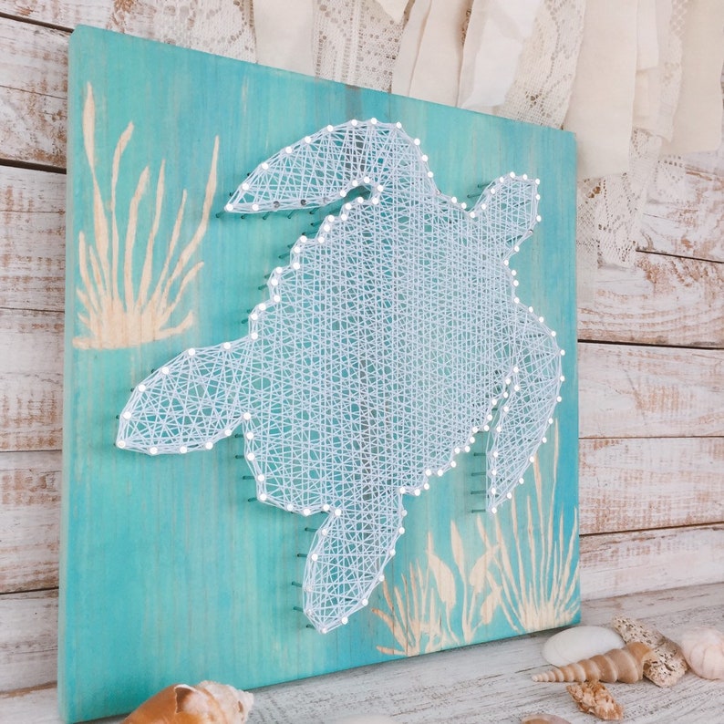 May include: A white string art sea turtle on a turquoise blue wooden background with a beige seagrass design.