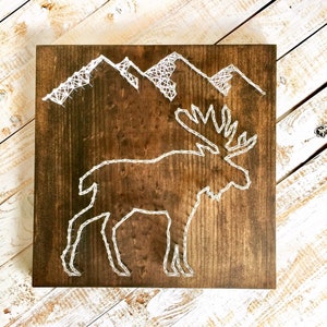 May include: A wooden wall hanging with a white string art design of a moose and mountains. The background is a dark brown wood.