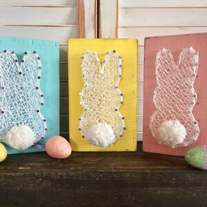 May include: Three wooden signs with string art of bunnies in the shape of an Easter egg. The signs are painted in pastel blue, yellow, and pink. Each sign has a white pom-pom for the bunny's tail.