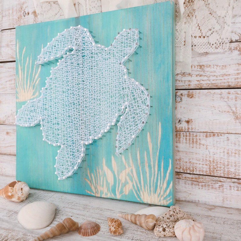 May include: A white string art sea turtle on a turquoise blue wooden background with a white woodgrain pattern. The turtle is surrounded by white string art seagrass.