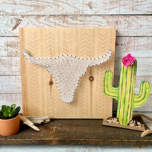 May include: A wooden wall art piece with a white string art design of a bull's head. The background is a light wood with a chevron pattern. A wooden cactus with a pink flower is on the right side of the image.