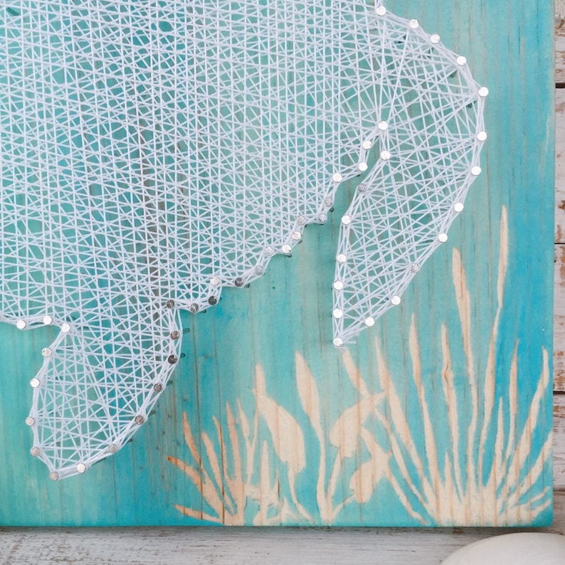 May include: White string art of a seahorse on a turquoise blue wooden background with a white stencil of seagrass in the bottom right corner.