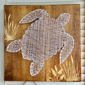 May include: White string art of a turtle on a brown wooden background. The turtle is surrounded by yellow painted seagrass.