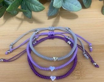 May include: Three friendship bracelets with silver heart charms. The bracelets are layered in shades of purple and gray.