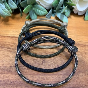 May include: Three braided rope bracelets in black, green, and camouflage patterns. The bracelets are tied in a knot and are stacked on top of each other.