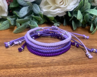 May include: Four handmade macrame bracelets in shades of purple, from light lavender to deep violet, are stacked on a wooden surface. Each bracelet features a sliding knot closure and silver-colored beads. The background includes white flowers and green foliage.