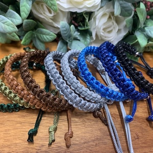 May include: A set of eight braided bracelets in various colours, including green, brown, grey, blue, and black. The bracelets are made of a woven cord material and have adjustable closures.