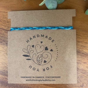 Può includere: Un braccialetto blu intrecciato è appoggiato su un cartoncino marrone. Il cartoncino presenta le parole "HANDMADE FOR YOU" in un design circolare con cuori e foglie. È visibile anche il testo "HANDMADE IN CANNOCK, STAFFORDSHIRE".