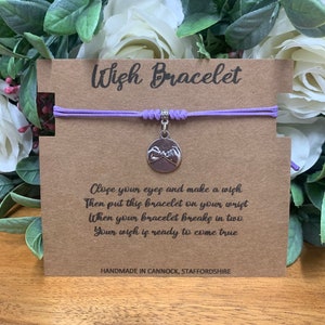 May include: A purple wish bracelet with a silver charm featuring two hands making a pinky promise. The card reads "Wish Bracelet" and includes instructions on how to make a wish. "Close your eyes and make a wish. Then put this bracelet on your wrist. When your bracelet breaks in two, your wish is ready to come true." The card also states "Handmade in Cannock, Staffordshire."