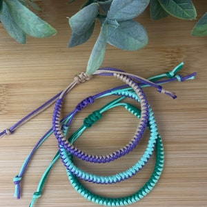 May include: Three braided friendship bracelets in green, purple and brown colours. The bracelets are knotted together and laid out on a wooden surface.