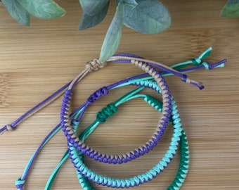 May include: Three braided friendship bracelets in green, purple, and brown colors. The bracelets are knotted together and laid out on a wooden surface.