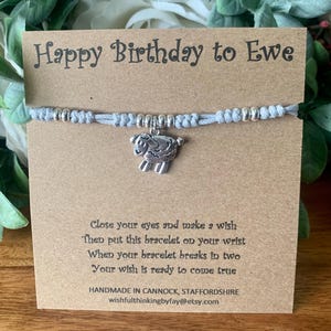 Könnte beinhalten: Ein graues Armband aus Schnur mit einem silbernen Anhänger in Form eines Schafs. Das Armband befindet sich auf einer braunen Karte mit dem Text "Happy Birthday to Ewe" und Anweisungen, wie man einen Wunsch macht.