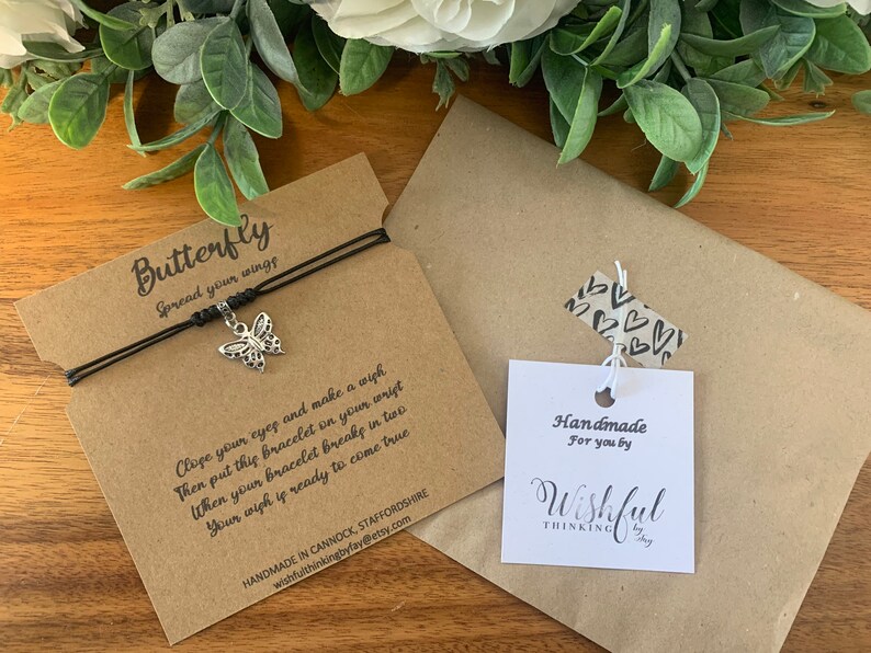May include: A black string bracelet with a silver butterfly charm. The bracelet is packaged on a brown card with the text "Butterfly Spread your wings" and instructions on how to make a wish. The card also includes the text "Handmade in Cannock, Staffordshire wishfulthinkingbyfay@etsy.com". A separate white card with the text "Handmade for you by Wishful Thinking by Fay" is included.