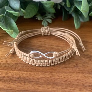 May include: Two tan macrame bracelets with a silver infinity symbol charm. One bracelet is woven, the other is multiple cords. The bracelets are on a wooden surface.