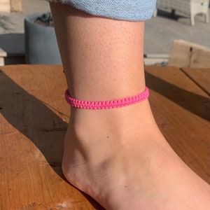 May include: A bright pink macrame anklet is worn on an ankle. The anklet is made of woven cord and is a vibrant, eye-catching accessory. The ankle is next to a wooden surface and a person's blue jeans.