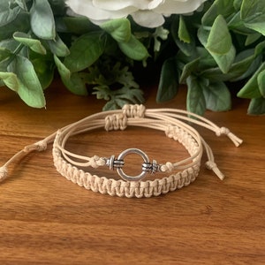 May include: A double-layered beige macrame bracelet with a silver circle charm. The bracelet is made with a knotting technique and has a simple, minimalist design.