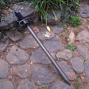 May include: A long metal sword featuring a dark, textured handle with a black wrap and a prominent metal hand guard. The blade is straight, double-edged, and tapers to a sharp point. The sword has a metallic finish with visible signs of wear.