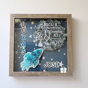 Op de afbeelding: Een ingelijste schaduwdoos met een blauwe en witte raket, sterren en een bloemenkrans. De krans heeft de tekst "March 8, Two Thousand Twenty Two, Livingston, NJ, 8:13 a.m., 7 lbs 19.75 oz" en de naam "Lukas" is verticaal in kleurrijke letters gespeld. De naam "Jose" staat in kleurrijke letters onderaan de doos.