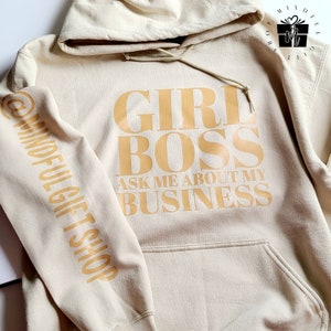 Peut inclure: Un sweat à capuche beige avec le texte "Girl Boss Ask Me About My Business" imprimé en marron sur le devant. La manche gauche porte le texte "Mindful Gift Shop" imprimé en marron.
