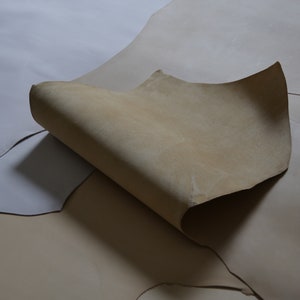 May include: Two pieces of light brown leather, one folded over the other. The leather is smooth and has a natural, slightly textured surface.