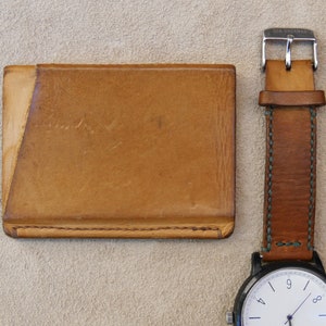 May include: Three leather wallets, one brown, one tan, and one light brown, with a brown leather watch strap and a white watch face with black hands.