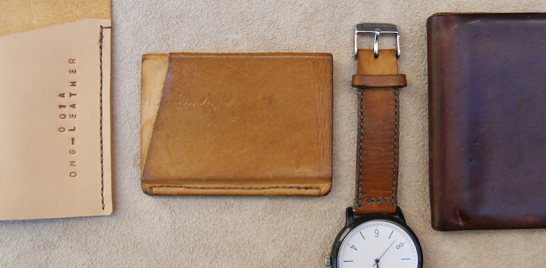 May include: Three leather wallets, one brown, one tan, and one light brown, with a brown leather watch strap and a white watch face with black hands.