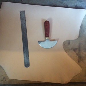 May include: A piece of light brown leather on a wooden surface with a metal ruler and a leather working tool with a wooden handle.