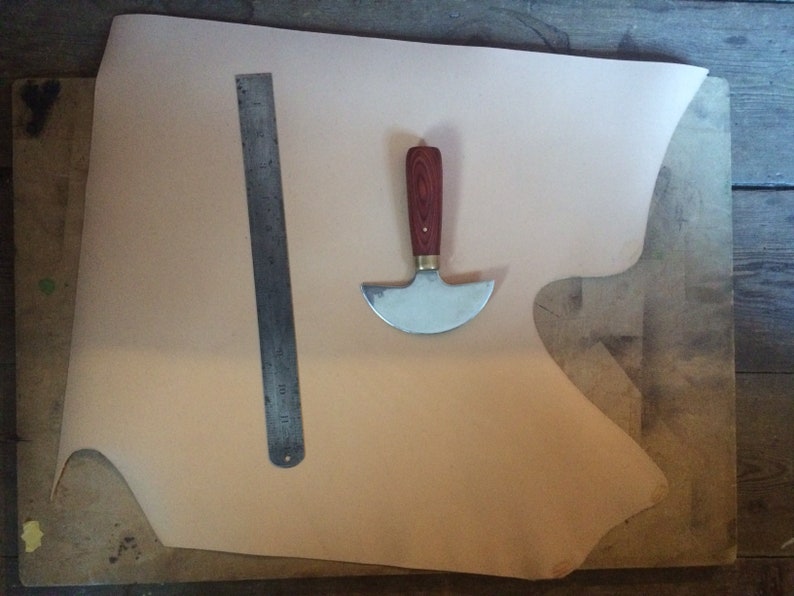 May include: A piece of light brown leather on a wooden surface with a metal ruler and a leather working tool with a wooden handle.
