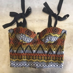 May include: A colorful bustier top with black tie straps. The top features a geometric pattern in orange, yellow, white, and black. The bustier has a sweetheart neckline and is made of woven fabric.