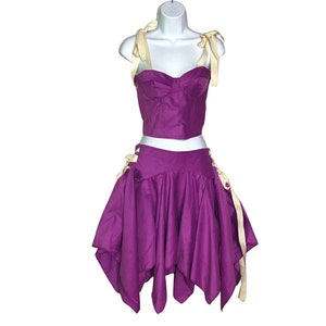 May include: A two-piece purple outfit on a mannequin. The top is a bustier with cream-colored straps. The skirt is a handkerchief style with cream-colored ties at the sides. The outfit is made of a shiny fabric.