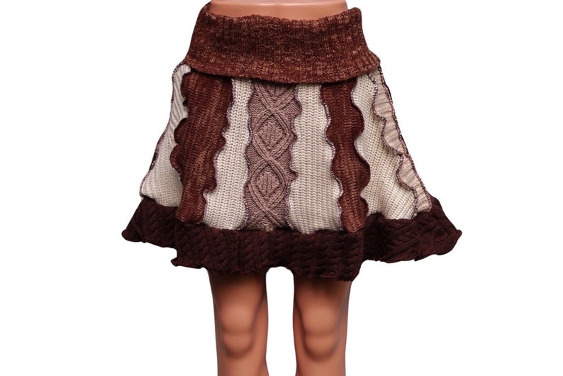 May include: A brown and cream-colored knitted skirt with a unique design. The skirt features a high, ribbed waistband and a flared hem. The skirt has a mix of textures, including cable knit and wavy patterns. The skirt is displayed on a mannequin.