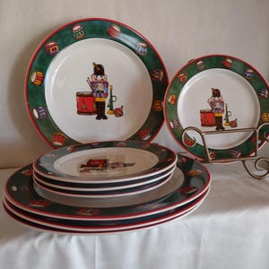 May include: Set of six Christmas themed dinner plates featuring a nutcracker design with red, green, and white colors. The plates are stacked on a white tablecloth.