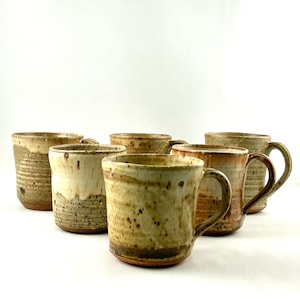 Peut inclure: Ensemble de sept mugs en céramique faits à la main. Les mugs présentent un aspect rustique et moucheté avec des tons bruns, beiges et verts. Chaque mug est doté d'une anse et d'une forme organique unique, pour le café ou le thé.