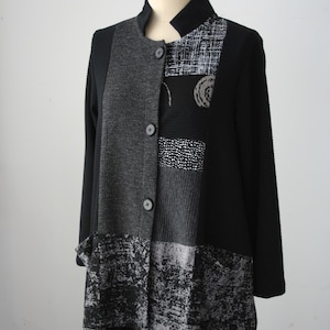 May include: A black and gray patchwork jacket with a mandarin collar and long sleeves. The jacket features a mix of textures and patterns, including solid black, textured gray, and patterned panels. It has three buttons down the front and two pockets.