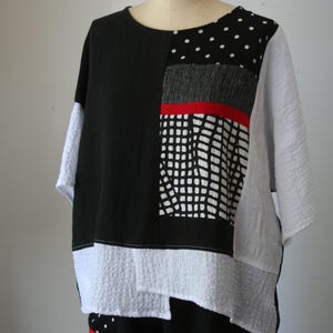 May include: A patchwork top in black, white, and gray, displayed on a mannequin. The top features a mix of textures and patterns, including polka dots, a grid design, and a red stripe. The sleeves are short and white.