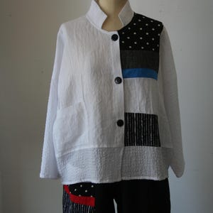 May include: White jacket with a mandarin collar and black buttons. The jacket features patchwork details with polka dots, stripes, and a blue accent. The bottom half of the jacket has a textured fabric. The jacket is paired with black pants.