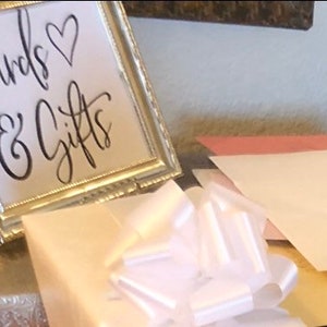 May include: A framed sign with the words "Cards & Gifts" in elegant script, accompanied by a heart symbol. A white bow and several envelopes are in the foreground, suggesting a celebratory event.