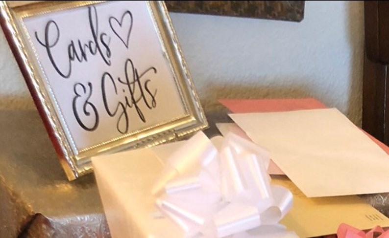 May include: A framed sign with the words "Cards & Gifts" in elegant script, accompanied by a heart symbol. A white bow and several envelopes are in the foreground, suggesting a celebratory event.